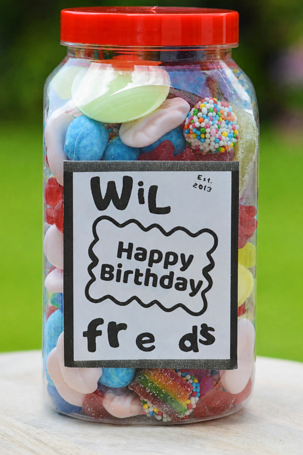 A clear plastic jar filled with assorted colorful gummy candies and sweets, topped with a bright red lid. A white label on the front reads 'Wil Happy Birthday freds Est.