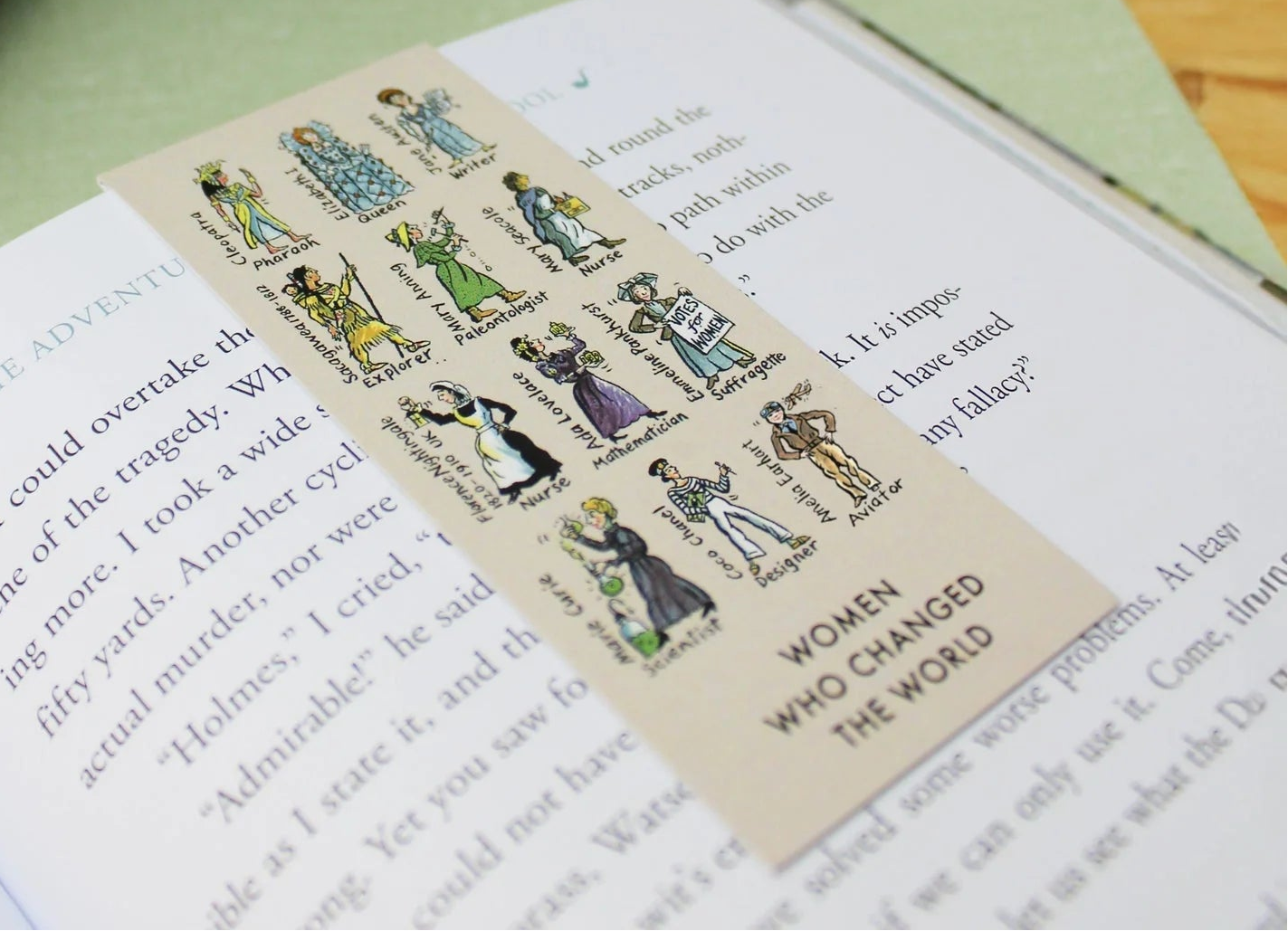 Open book with a bookmark featuring illustrations of women, next to a mug on a table.