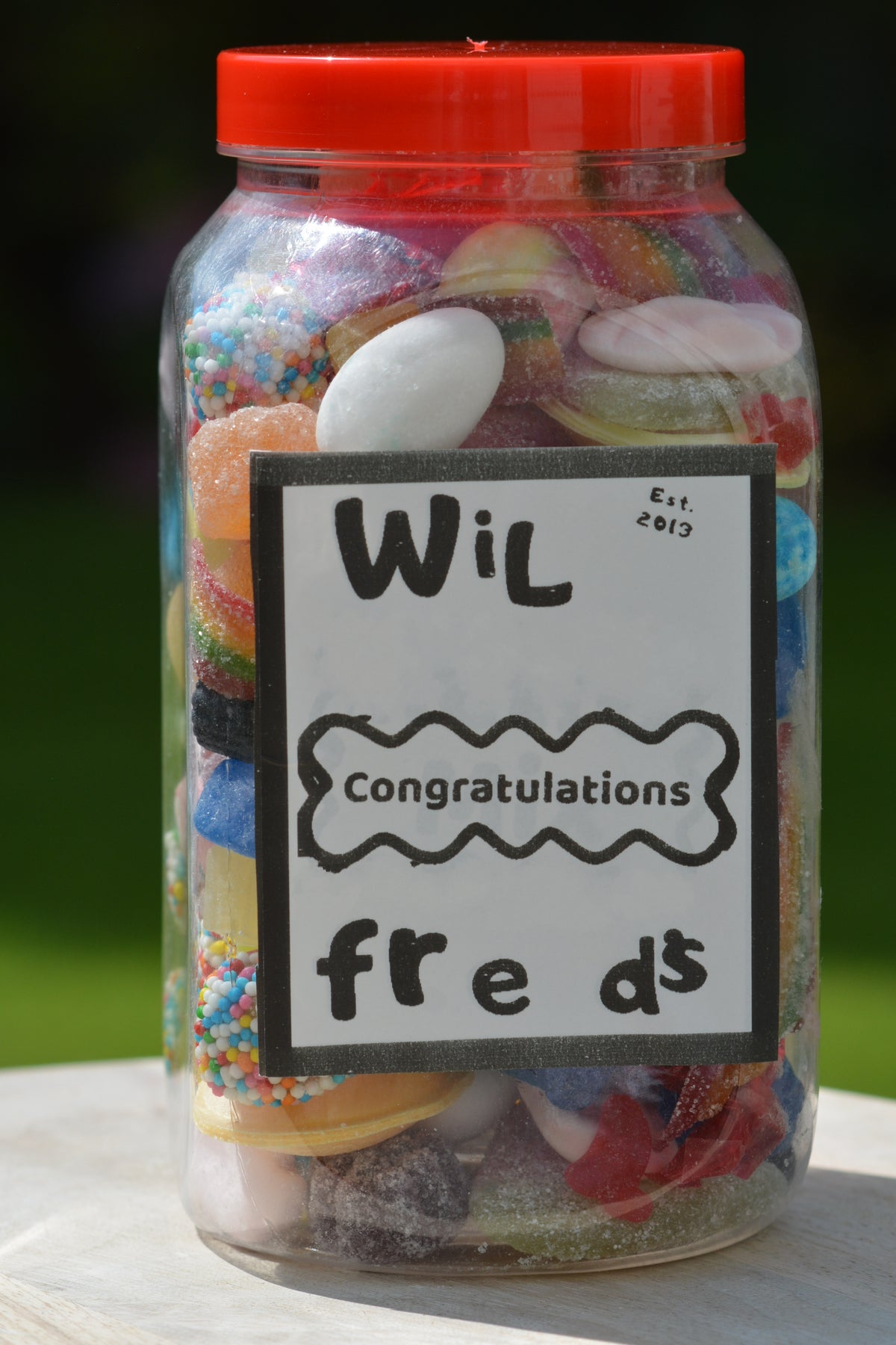 A clear plastic jar filled with assorted colorful candies and sweets, topped with a bright red lid. A white label on the front reads 'Wilfred Congratulations freds' in black handwritten-style text.