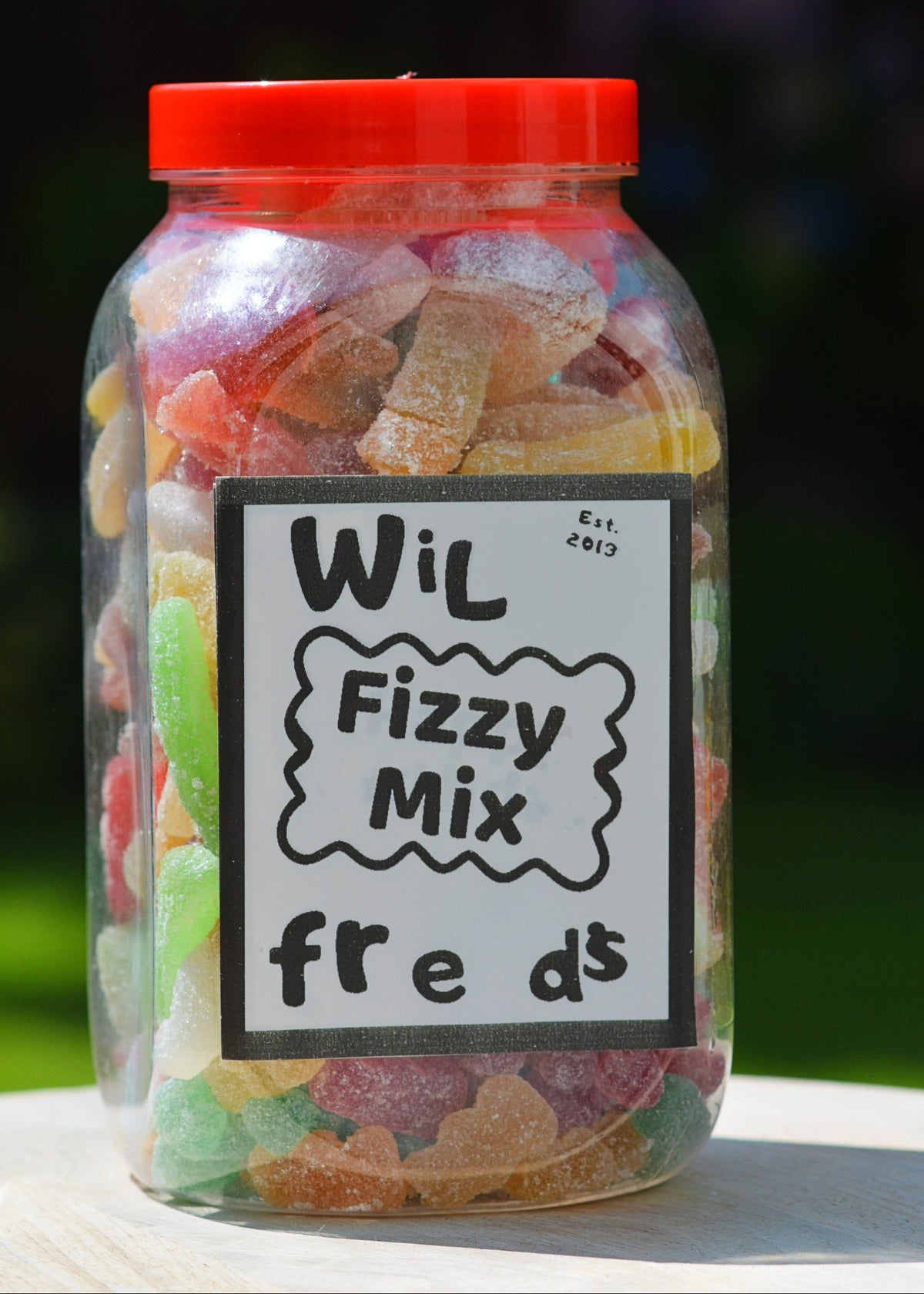 A clear plastic jar filled with assorted colorful gummy candies, topped with a bright red lid and labeled 'Wil Fizzy Mix freds' on a white background with a blurred green outdoor setting behind it.
