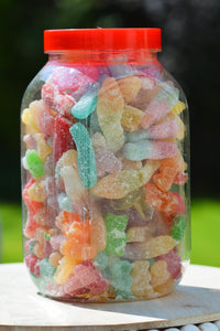A clear plastic jar filled with assorted colorful gummy candies and topped with a bright red lid, photographed outdoors on a light surface with a blurred green background.
