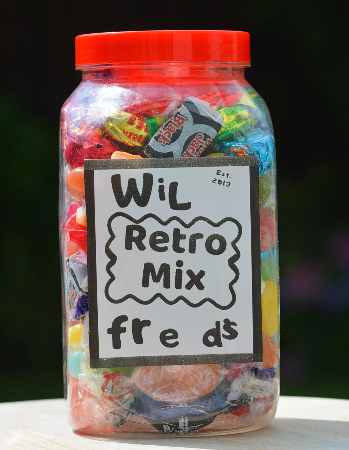 A clear plastic jar filled with assorted colorful wrapped candies, topped with a bright red lid and labeled 'Wil Retro Mix freds' on a white sign, set on a light wooden surface with a blurred dark green background.