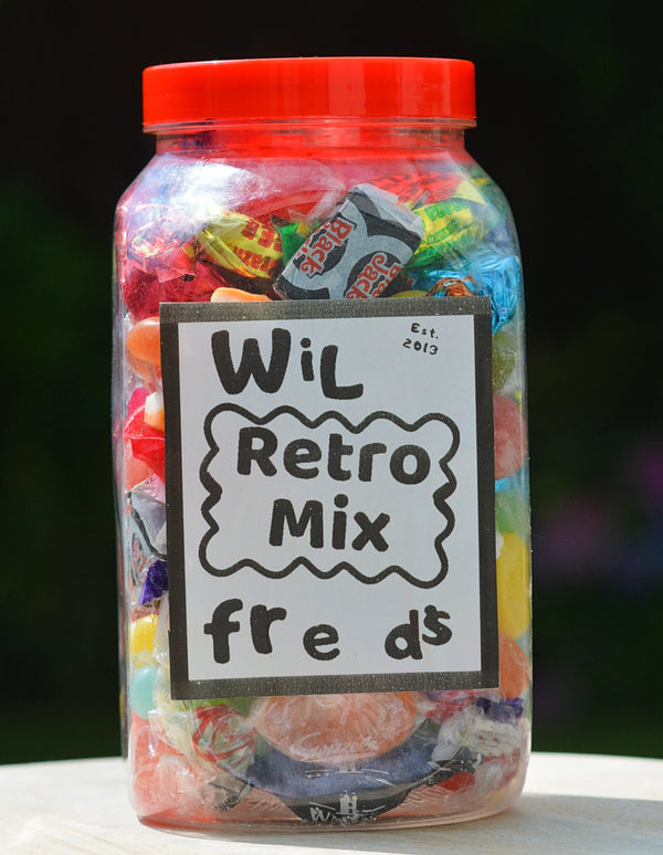 A clear plastic jar filled with assorted colorful wrapped candies, topped with a bright red lid and labeled 'Wil Retro Mix freds' on a white sign, set on a light wooden surface with a blurred dark green background.