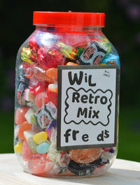 A clear plastic jar filled with assorted colorful retro candies and wrapped sweets, topped with a bright orange-red lid and labeled 'Wil Retro Mix freds' on a white sign, set on a light wooden surface outdoors with a blurred green background.