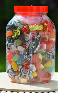 A large clear plastic jar filled with assorted colorful wrapped candies and gummy sweets, topped with a bright red lid and sitting on a light wooden surface outdoors.