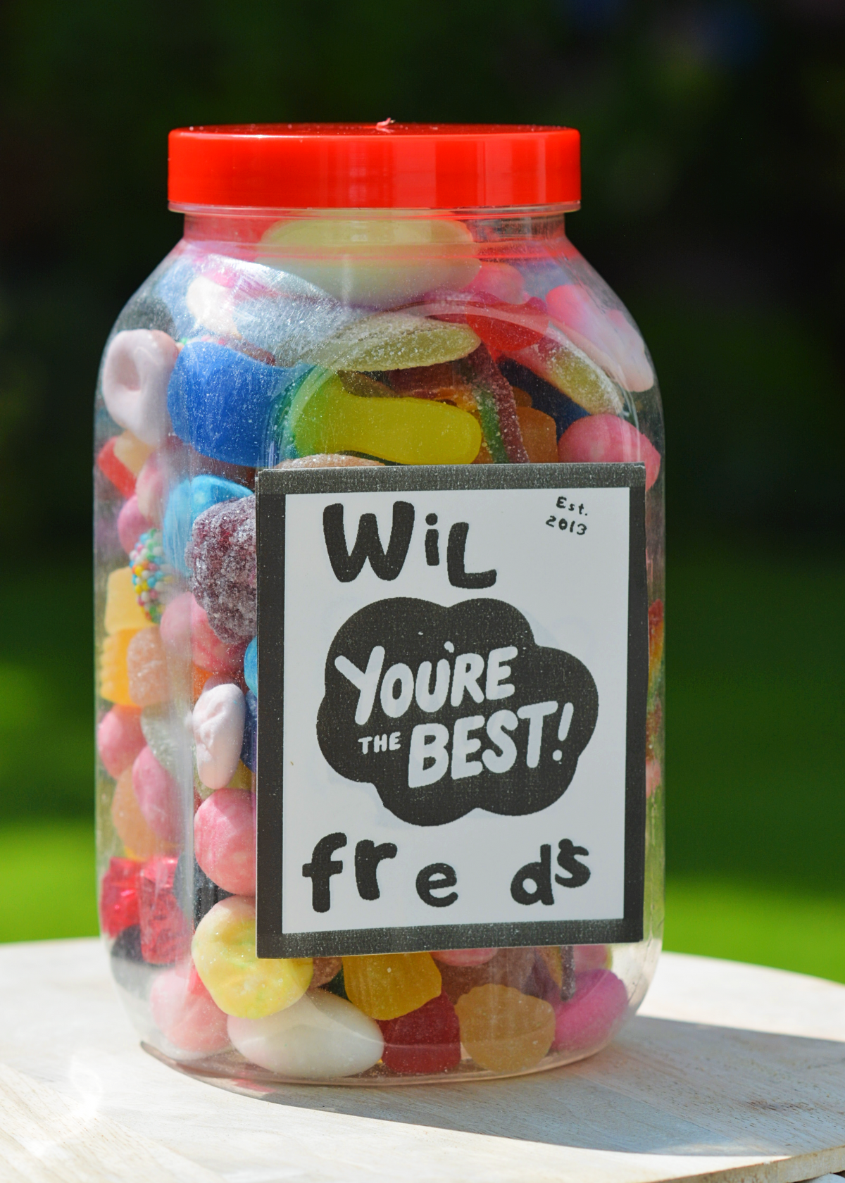 A clear plastic jar filled with assorted colorful gummy candies, topped with a bright red lid and featuring a black-and-white handwritten label that reads 'Wil You're the BEST! fre ds'. The jar is placed on a light wooden surface with a blurred green outdoor background.